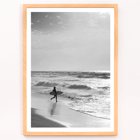 Surfer with a board by the ocean in a black and white picture titled Surfer in Portugal by artist Black Black and white poster Surfer in Portugal by Black & White Picture depicts a lone surfer entering the ocean carrying a surfboard.