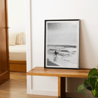 Surfer with a board by the ocean in a black and white picture titled Surfer in Portugal by artist Black Black and white poster Surfer in Portugal by Black & White Picture depicts a lone surfer entering the ocean carrying a surfboard.