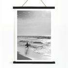 Surfer with a board by the ocean in a black and white picture titled Surfer in Portugal by artist Black Black and white poster Surfer in Portugal by Black & White Picture depicts a lone surfer entering the ocean carrying a surfboard.