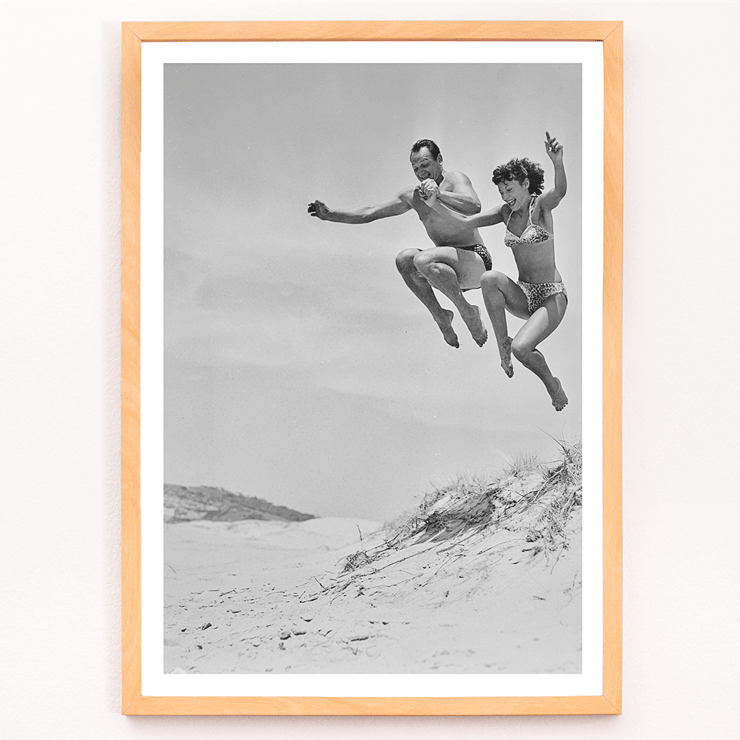 Acrobatic jump at the beach by Tivoli stars Acrobatics at the Beach Poster of Acrobatics at the Beach by Two Tivoli Stars featuring two people in swimsuits jumping on a sandy beach.