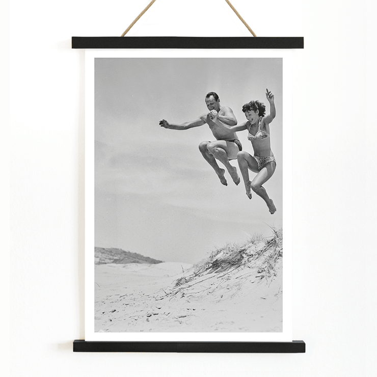 Acrobatic jump at the beach by Tivoli stars Acrobatics at the Beach Poster of Acrobatics at the Beach by Two Tivoli Stars featuring two people in swimsuits jumping on a sandy beach.