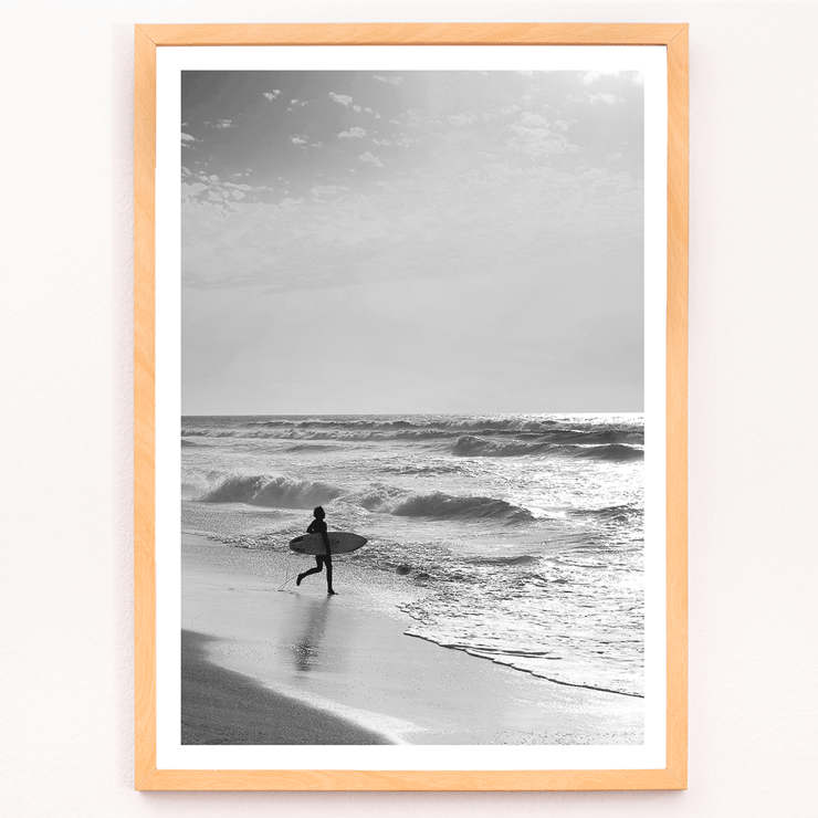 Surfer with a board by the ocean in a black and white picture titled Surfer in Portugal by artist Black Black and white poster Surfer in Portugal by Black & White Picture depicts a lone surfer entering the ocean carrying a surfboard.