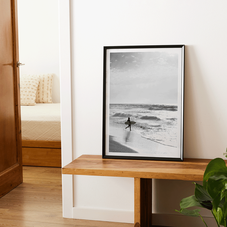 Surfer with a board by the ocean in a black and white picture titled Surfer in Portugal by artist Black Black and white poster Surfer in Portugal by Black & White Picture depicts a lone surfer entering the ocean carrying a surfboard.