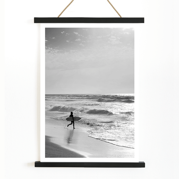 Surfer with a board by the ocean in a black and white picture titled Surfer in Portugal by artist Black Black and white poster Surfer in Portugal by Black & White Picture depicts a lone surfer entering the ocean carrying a surfboard.